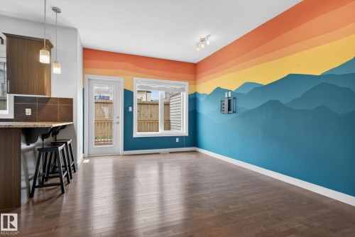 Open concept living area featuring wood-finish flooring, a vibrant accent wall with a mountain mural, a large window, and a glass-paneled door leading to the exterior - 11 450 Mcconachie Way, Edmonton, AB - Indoor Photo Showing Other Room