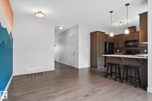 Open-concept living space featuring wood-finish flooring, a kitchen island with pendant lighting, dark wood cabinetry, and integrated black appliances - 11 450 Mcconachie Way, Edmonton, AB - Indoor Photo Showing Kitchen