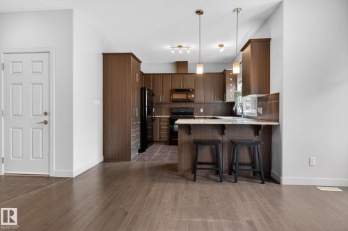 Open-concept kitchen featuring wood-finish cabinetry, black appliances, and a breakfast bar with pendant lighting - 11 450 Mcconachie Way, Edmonton, AB - Indoor Photo Showing Kitchen