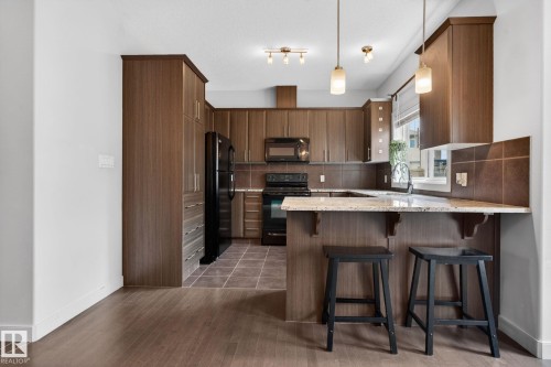 Modern kitchen featuring abundant wood-finish cabinetry, black appliances, a stone-finish countertop, and a tiled backsplash - 11 450 Mcconachie Way, Edmonton, AB - Indoor Photo Showing Kitchen With Upgraded Kitchen