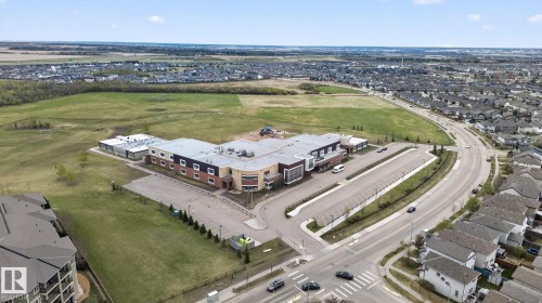 Aerial perspective showcasing a community building with a substantial parking area, adjacent to expansive green fields and residential rooftops - 11 450 Mcconachie Way, Edmonton, AB - Outdoor With View