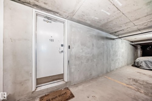 Concrete-walled parking area featuring a white entry door with number 105, a door closer, and a metal kick plate - 105 13825 155 Avenue, Edmonton, AB - Indoor