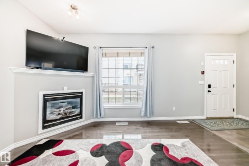 Corner-mounted electric fireplace with a white mantel, dark wood-finish flooring, a multi-pane window, white baseboards, and a paneled entry door - 105 13825 155 Avenue, Edmonton, AB - Indoor Photo Showing Living Room With Fireplace