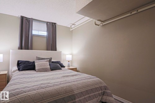 Neutral-toned room featuring a window with blinds and dark gray curtains, a white upholstered headboard, and light wood-finish nightstands with lamps - 1214 85 Street Nw, Edmonton, AB - Indoor Photo Showing Bedroom