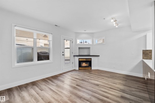 Spacious room featuring wood-finish flooring, a built-in fireplace with a dark mantel, multiple windows, and a glass-paned door - 205 10939 82 Avenue, Edmonton, AB - Indoor Photo Showing Living Room With Fireplace