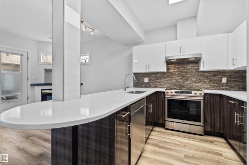 Contemporary kitchen featuring an expansive countertop island with an integrated sink and stainless steel faucet, two-tone cabinetry, a tile backsplash, and stainless steel appliances - 205 10939 82 Avenue, Edmonton, AB - Indoor Photo Showing Kitchen With Upgraded Kitchen