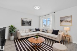 Living area featuring wood-finish flooring, a contemporary ceiling light fixture, a window with white curtains, and a black wrought iron railing - 