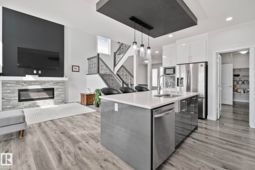 Open-concept living space featuring wood-finish flooring, a contemporary kitchen island with a white countertop and pendant lighting, and a stacked stone fireplace - 334 Aston Close, Leduc, AB - Indoor Photo Showing Kitchen With Fireplace With Upgraded Kitchen
