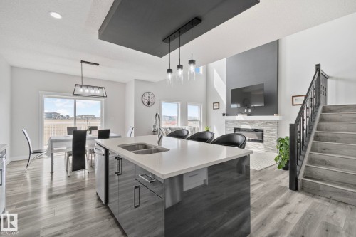 Open-concept interior featuring a kitchen island with a double sink and white countertop, wood-finish flooring, a contemporary fireplace with a stone surround, a prominent staircase with dark railings, and sliding glass doors leading to an exterior deck - 334 Aston Close, Leduc, AB - Indoor Photo Showing Kitchen With Double Sink With Upgraded Kitchen