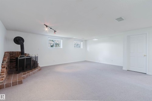 Spacious room featuring a brick hearth and wood-burning stove, light-colored carpeting, white walls, two windows, and track lighting - 13755 131B Avenue, Edmonton, AB - Indoor Photo Showing Other Room With Fireplace