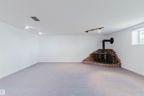 Spacious room featuring a brick wood stove with a black stovepipe, track lighting, recessed lighting, a white framed window, and light-colored carpeting - 13755 131B Avenue, Edmonton, AB - Indoor Photo Showing Other Room