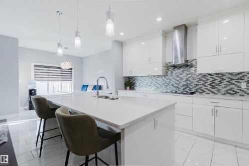 Contemporary kitchen featuring a large center island with integrated sink, white flat-panel cabinetry, a tile backsplash with a mosaic pattern, stainless steel range hood, and white tile flooring - 17030 62 Street, Edmonton, AB - Indoor Photo Showing Kitchen With Double Sink With Upgraded Kitchen