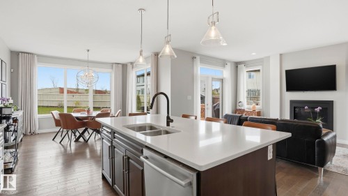 Open-concept main living area featuring a kitchen island with integrated sink, wood-finish flooring, a dining area with a large window, a living area with a fireplace, and a glass-paned door - 2620 Adam Crest, Edmonton, AB - Indoor Photo Showing Kitchen With Fireplace With Double Sink