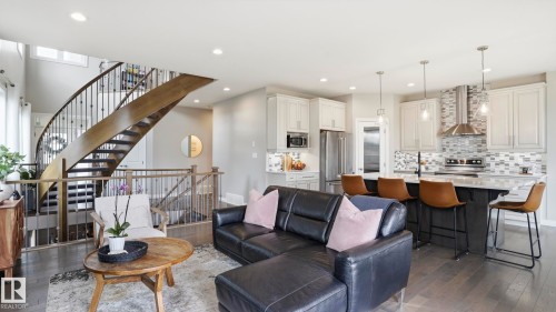 Open-concept living space featuring wood-finish flooring, a contemporary kitchen with stainless steel appliances, a large central island with seating, a mosaic tile backsplash, and a modern curved staircase with wrought iron balusters - 2620 Adam Crest, Edmonton, AB - Indoor Photo Showing Living Room