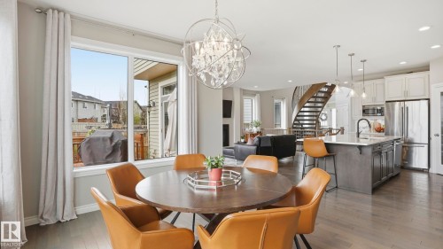 Spacious dining area with a contemporary chandelier, large window overlooking a rear deck, light wood-finish flooring, and an open-concept layout leading to a modern kitchen and a curved staircase - 2620 Adam Crest, Edmonton, AB - Indoor Photo Showing Dining Room