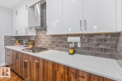 Kitchen featuring white upper cabinetry, wood-finish lower cabinetry, a light-toned countertop, a built-in gas cooktop, and a stainless steel range hood with glass accents - 219 Cranberry Bend, Fort Saskatchewan, AB - Indoor Photo Showing Kitchen With Upgraded Kitchen