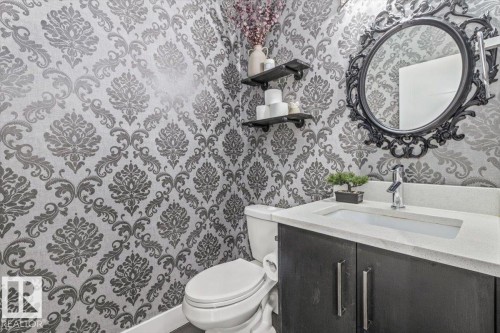 Powder room featuring ornamental wallpaper, a dark wood-finish vanity with a light countertop, an integrated sink basin, and a decorative black-framed mirror - 219 Cranberry Bend, Fort Saskatchewan, AB - Indoor Photo Showing Bathroom