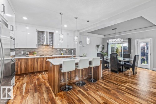 Modern kitchen featuring wood-finish flooring, white cabinetry, a large kitchen island with seating, stainless steel appliances, and a tiled backsplash - 219 Cranberry Bend, Fort Saskatchewan, AB - Indoor