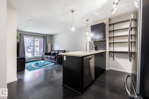 Open-concept living space featuring dark wood-finish flooring, a kitchen island with a light-toned countertop, stainless steel appliances, and built-in shelving - 131 344 Windermere Road, Edmonton, AB - Indoor Photo Showing Kitchen With Upgraded Kitchen