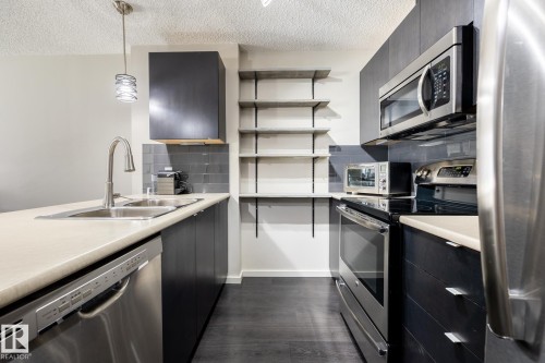 Modern kitchen featuring dark cabinetry, stainless steel appliances, light countertops, a double basin sink with a gooseneck faucet, and open shelving - 131 344 Windermere Road, Edmonton, AB - Indoor Photo Showing Kitchen With Double Sink