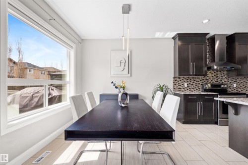 Open-concept dining area featuring a large window, contemporary linear pendant lighting, light wood-finish flooring, and dark cabinetry in the adjacent kitchen - 3807 Claxton Close, Edmonton, AB - Indoor