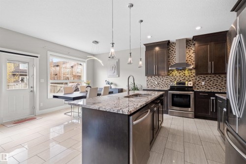 Spacious kitchen featuring an island with stone countertop, stainless steel appliances, dark wood cabinetry, a mosaic tile backsplash, and light tile flooring - 3807 Claxton Close, Edmonton, AB - Indoor Photo Showing Kitchen With Upgraded Kitchen