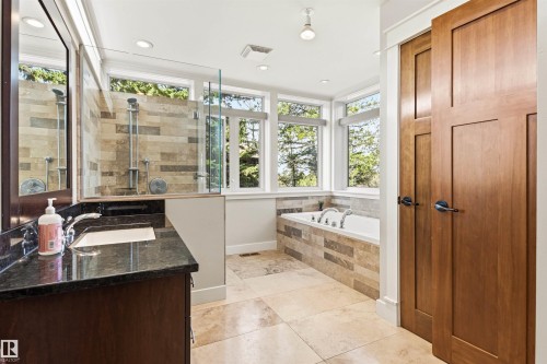 Contemporary bathroom featuring a large soaking tub with stone tile surround, a glass-enclosed shower with stone tile, a vanity with a dark countertop and undermount sink, and light wood-paneled doors - 9010 120 Street, Edmonton, AB - Indoor Photo Showing Bathroom