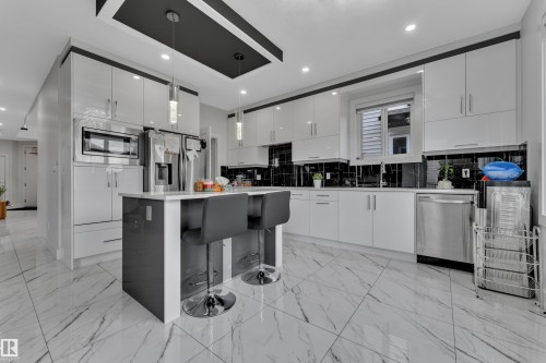 Modern kitchen featuring a central island with seating, white shaker-style cabinetry, stainless steel appliances, black tile backsplash, and large format marble-look floor tiles - 1827 22 Street Nw, Edmonton, AB - Indoor Photo Showing Kitchen With Upgraded Kitchen