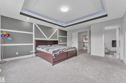 Spacious sleeping area featuring a tray ceiling with integrated lighting, recessed lighting fixtures, light gray carpet flooring, a decorative accent wall with white trim, and a doorway to a bathroom with tile flooring - 1827 22 Street Nw, Edmonton, AB - Indoor Photo Showing Bedroom