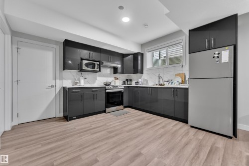 Kitchen featuring dark glossy cabinetry, light countertops, wood-finish flooring, stainless steel appliances, and a tiled backsplash - 1827 22 Street Nw, Edmonton, AB - Indoor Photo Showing Kitchen