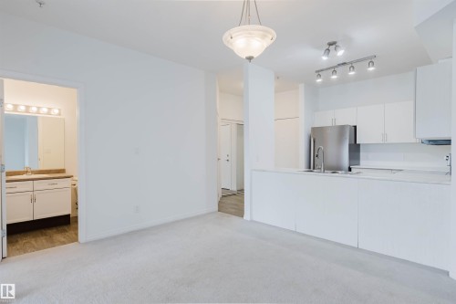 Open-concept living area with light-toned carpeting and a kitchen featuring white cabinetry, stainless steel appliances, and track lighting - 308 4316 139 Avenue, Edmonton, AB 