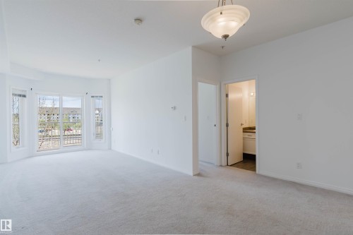 Spacious room featuring light-toned carpet flooring, a large bay window with multiple panes, and a contemporary ceiling light fixture - 308 4316 139 Avenue, Edmonton, AB 