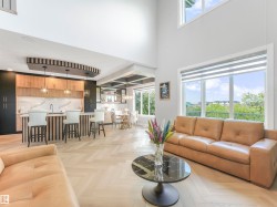 Open-concept living area featuring high ceilings, large windows, wood-finish herringbone flooring, a built-in kitchen island with pendant lighting, and a dining area with a coffered ceiling - 