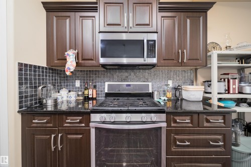 Kitchen featuring dark wood-finish cabinetry, stainless steel oven with gas range, stainless steel microwave, black countertops, and a mosaic tile backsplash - 1508 Cunningham Cape Cape, Edmonton, AB - Indoor Photo Showing Kitchen