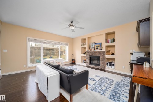 Living area featuring wood-finish flooring, a stone-clad fireplace, integrated shelving, a ceiling fan, and large windows - 1508 Cunningham Cape Cape, Edmonton, AB - Indoor Photo Showing Living Room With Fireplace
