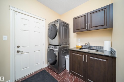 Laundry area featuring stacked appliances, dark wood-finish cabinetry, a black countertop, stainless steel sink, and a mosaic tile backsplash - 1508 Cunningham Cape Cape, Edmonton, AB - Indoor Photo Showing Laundry Room