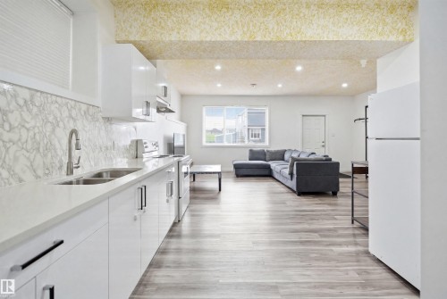 Basement kitchen featuring white cabinetry, a double basin stainless steel sink, light-colored countertops, and a marble-patterned backsplash - 3909 49 Ave, Beaumont, AB - Indoor Photo Showing Kitchen With Double Sink