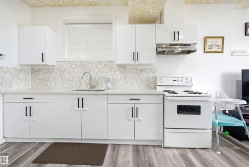 Bright kitchen featuring white cabinetry with black hardware, a stainless steel sink, a range with an overhead vent, a white countertop, and a marble-patterned backsplash - 3909 49 Ave, Beaumont, AB - Indoor Photo Showing Kitchen