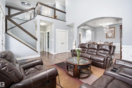 Open-concept living space featuring wood-finish flooring, a contemporary glass and wood railing staircase, arched doorways, tray ceiling with chandelier, and wainscoting - 3909 49 Ave, Beaumont, AB - Indoor Photo Showing Living Room