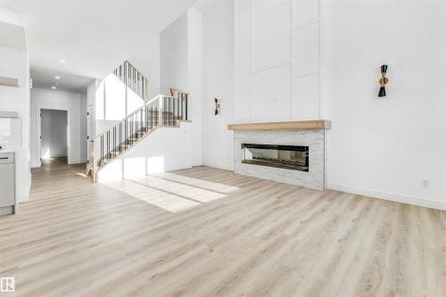 The property features light-toned flooring, a fireplace with a light-colored stone surround and a wooden mantle, and a staircase with light wood steps and dark metal railings - 4102 67 Street, Beaumont, AB - Indoor Photo Showing Living Room With Fireplace