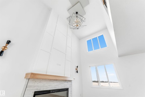 The property features a soaring ceiling with a contemporary chandelier, large windows providing natural light, and a fireplace with a light wood mantel and tiled surround - 4102 67 Street, Beaumont, AB - Indoor Photo Showing Other Room With Fireplace