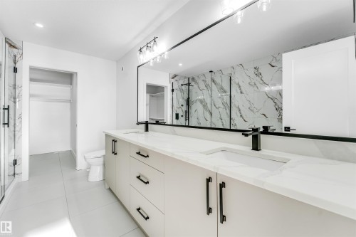 The bathroom features a double vanity with light-colored cabinetry and dark hardware, a large mirror, and a shower with a marble-patterned tiled wall - 4102 67 Street, Beaumont, AB - Indoor Photo Showing Bathroom