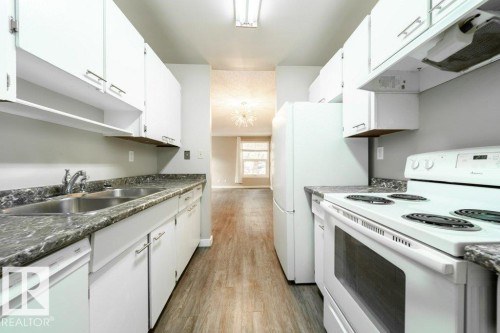 Galley kitchen featuring wood-finish flooring, white cabinetry, dark stone-look countertops, a double basin sink, and white appliances - 17106 108 St Nw, Edmonton, AB - Indoor Photo Showing Kitchen With Double Sink