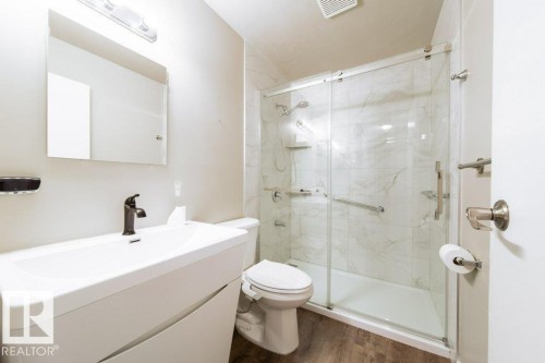 Contemporary bathroom featuring a white vanity with integrated sink, a rectangular mirror, a glass-enclosed shower with marble-patterned wall tiles, and wood-finish flooring - 17106 108 St Nw, Edmonton, AB - Indoor Photo Showing Bathroom