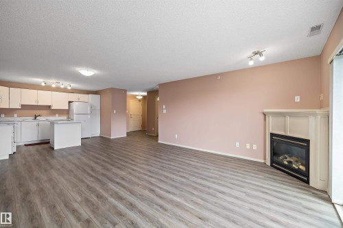 Open-concept living space featuring wood-finish flooring, a white kitchen with a peninsula, white appliances, and a corner-set fireplace with a light-toned mantel - 406 16035 132 Street, Edmonton, AB - Indoor With Fireplace