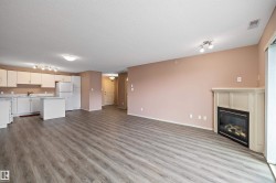 Open-concept living space featuring wood-finish flooring, a white kitchen with a peninsula, white appliances, and a corner-set fireplace with a light-toned mantel - 