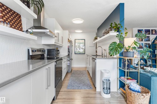 Modern galley kitchen featuring white cabinetry, light countertops, stainless steel appliances, a white geometric tile backsplash, and light wood-finish flooring - 217 14819 51 Avenue, Edmonton, AB - Indoor Photo Showing Kitchen