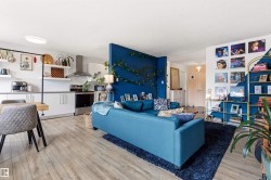 Open-concept living space featuring wood-finish flooring, a kitchen with stainless steel appliances, white cabinetry, and floating shelves - 