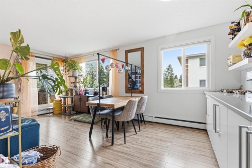 Open-concept living space with wood-finish flooring, a contemporary dining area, a large window, a glass-paned exterior door, and sleek white cabinetry with modern hardware - 217 14819 51 Avenue, Edmonton, AB - Indoor