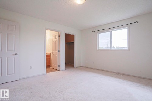 Carpeted room featuring a double-pane window with a decorative curtain rod, a ceiling-mounted light fixture, and two panel doors, one revealing a bathroom vanity and the other a closet with a hanging rod - 1627 Melrose Place Sw, Edmonton, AB - Indoor